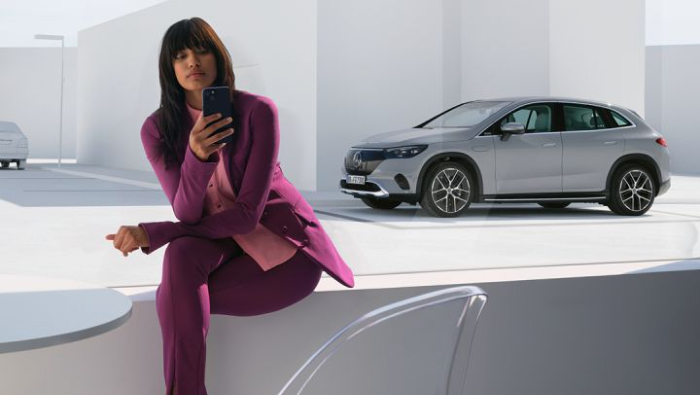 Eine Dame sitzt mit ihrem Handy und macht sich online ihren Termin aus, im Hintergrund ein grauer Mercedes