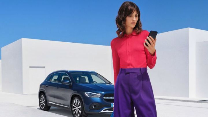 Eine Dame steht mit ihrem Handy und im Hintergrund ein blauer Mercedes